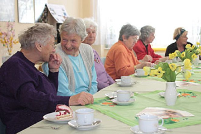 Foto: Zwei ältere Damen unterhalten sich beschwingt bei Kaffee und Kuchen. Sie sitzen an einem langen, gedeckten Tisch mit gelben Tulpen. Um sie herum sitzen auch andere Gäste.